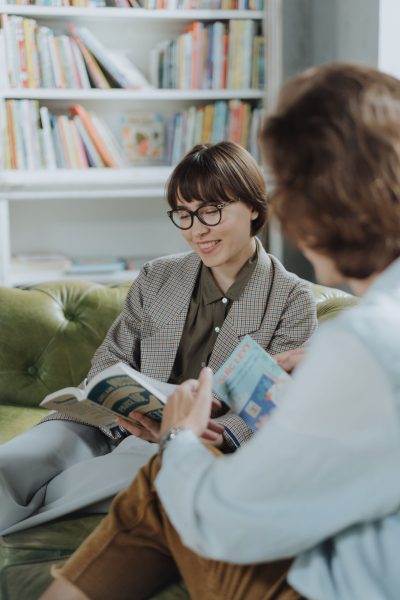 Two young adults enjoy reading on a sofa in a cozy library setting, surrounded by books.